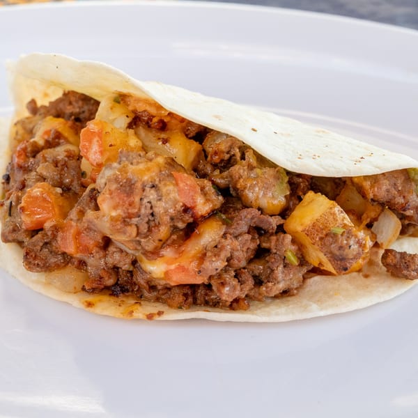 A Super Burrito taco filled with seasoned meat, potatoes, tomatoes, and melted cheese on a white plate.