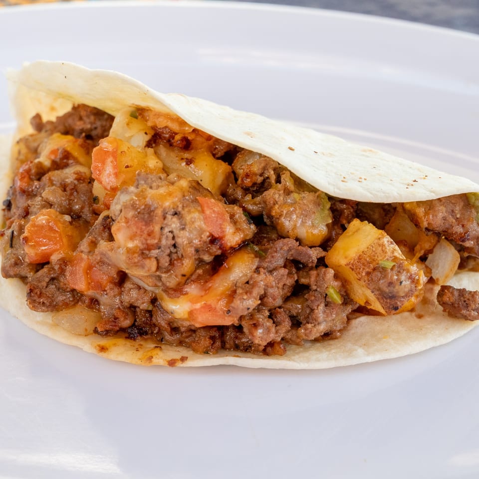 A Super Burrito taco filled with seasoned meat, potatoes, tomatoes, and melted cheese on a white plate.