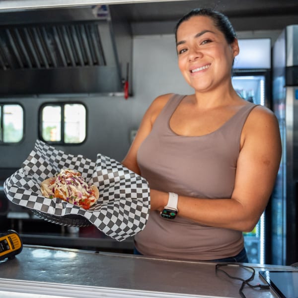 A smiling woman in a food truck holds a Super Burrito hot dog in checkered paper.
