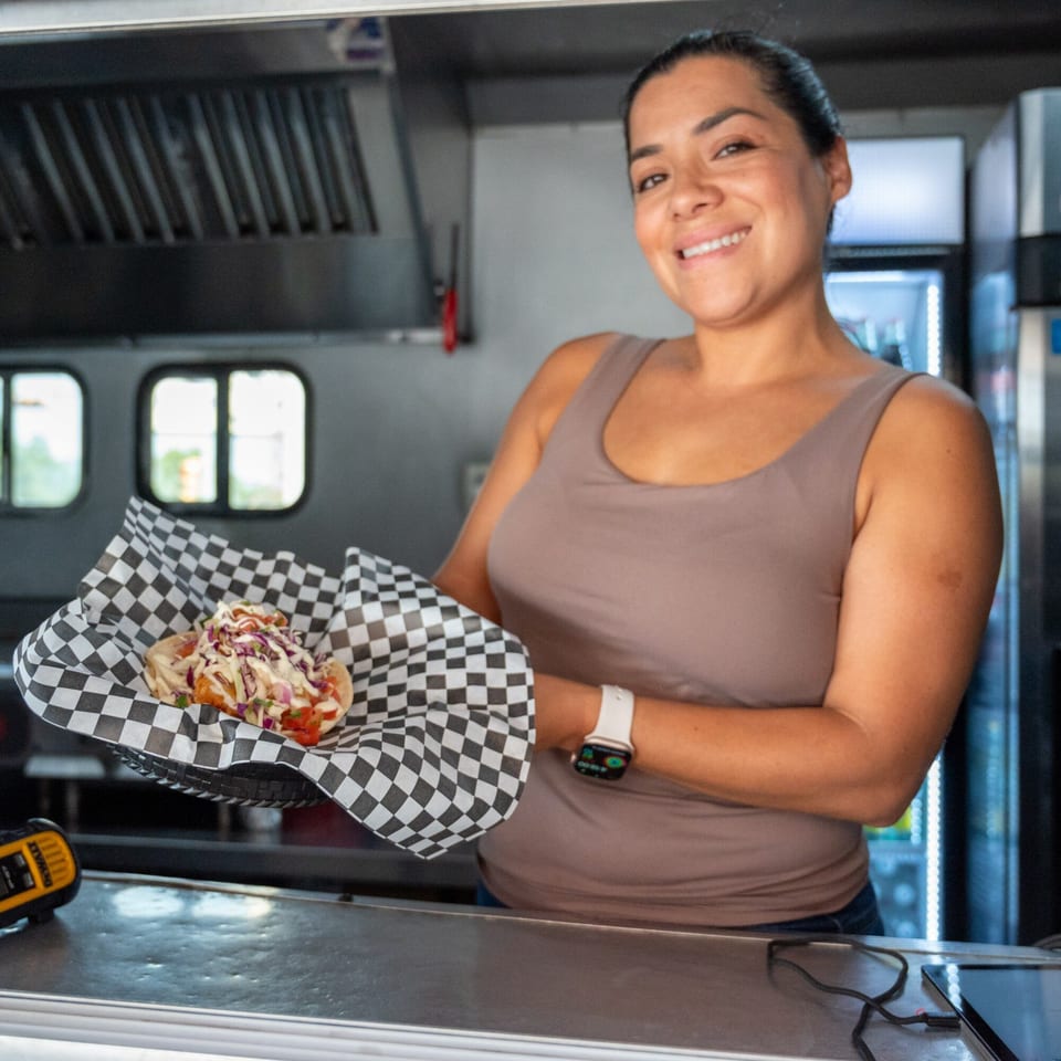 A smiling woman in a food truck holds a Super Burrito hot dog in checkered paper.