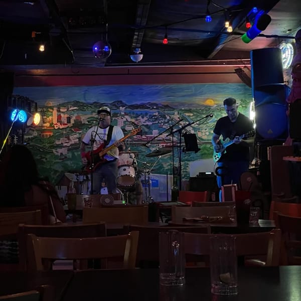 A band performing on stage with a colorful mural backdrop, viewed from a dark dining area.