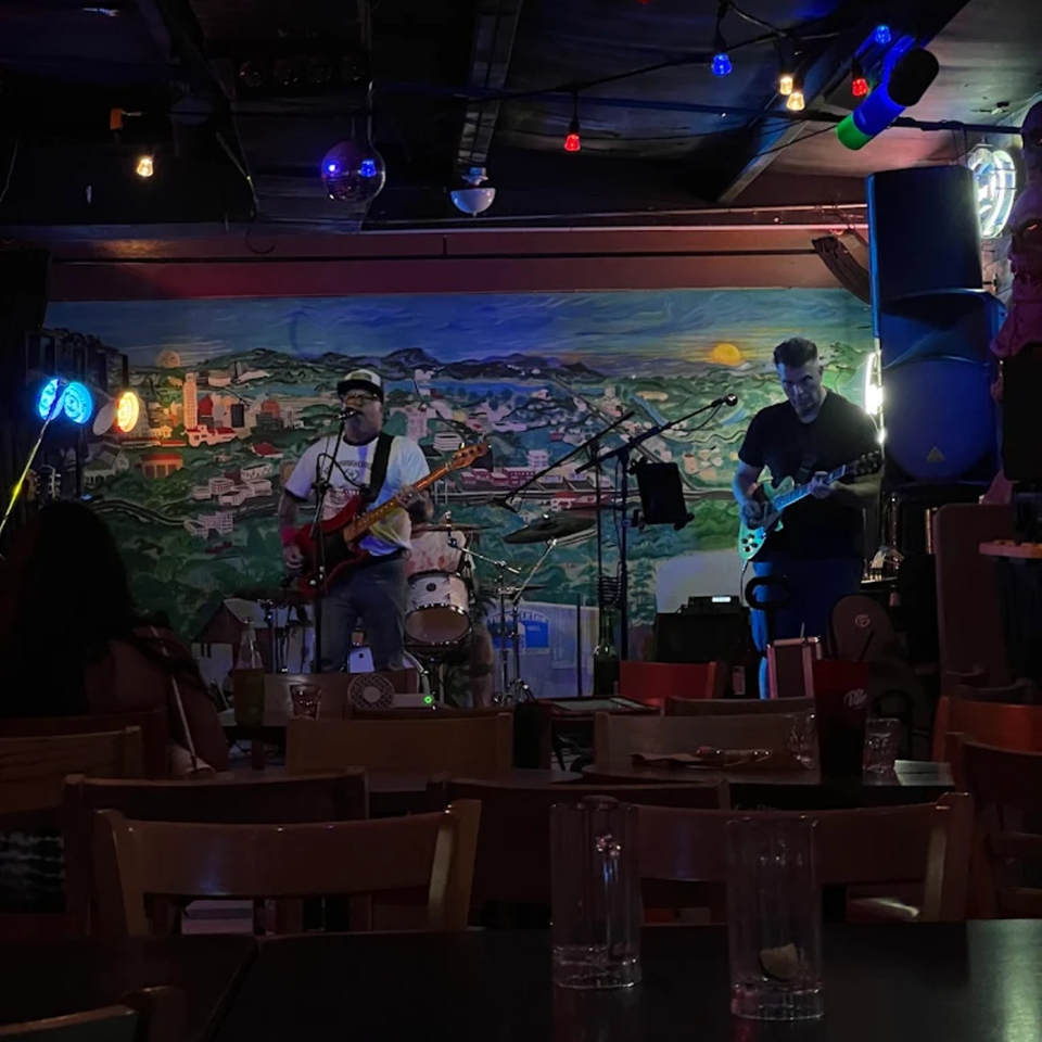 A band performing on stage with a colorful mural backdrop, viewed from a dark dining area.