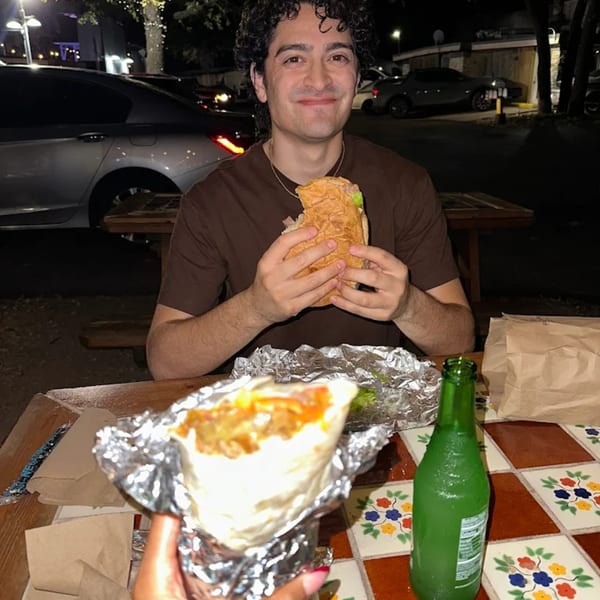 A man smiles while holding a large Super Burrito at an outdoor picnic table at night.
