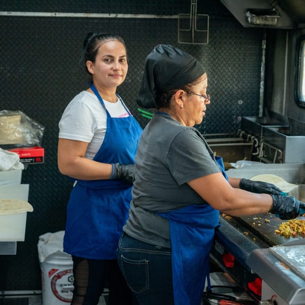 Two women in blue aprons preparing Mexican food on a griddle inside a Super Burrito food truck.