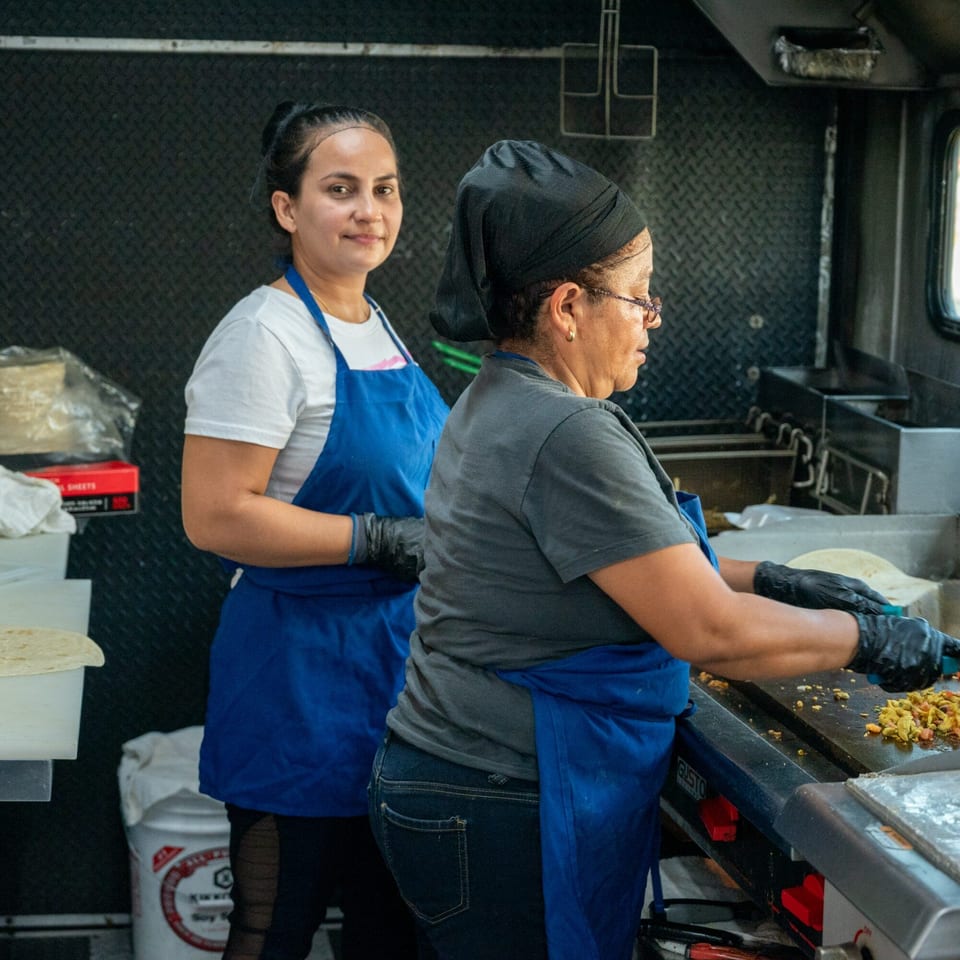 Two women in blue aprons preparing Mexican food on a griddle inside a Super Burrito food truck.