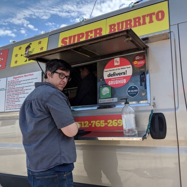 A man in glasses stands in front of the Super Burrito food truck, which has a menu and delivery stickers.