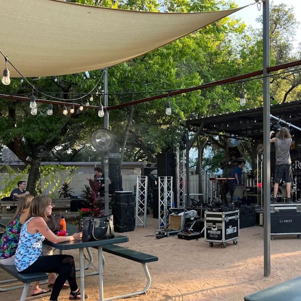 Patrons seated at picnic tables under a canopy, watching a band set up on an outdoor stage.