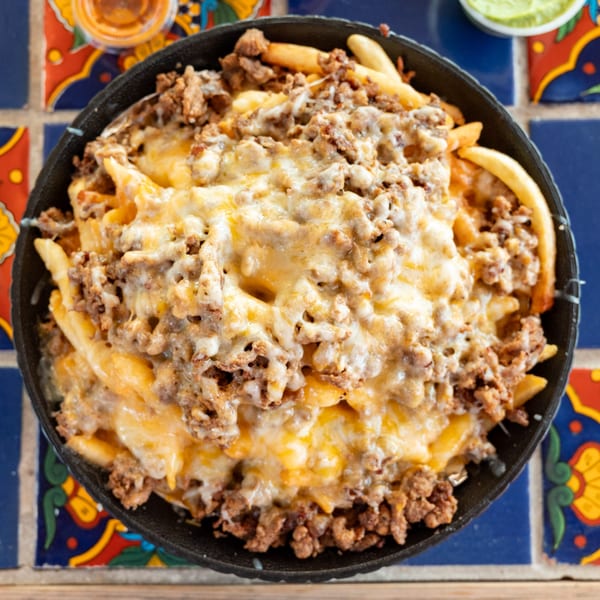 A black skillet filled with golden french fries, ground beef, and melted cheese on a colorful tiled table.